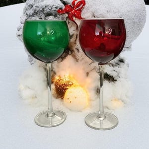 May include: Two stemmed wine glasses, one green and one red, stand in the snow. The glasses have a rounded bowl and a long, slender stem. A red bow is in the background, with a snowy tree and a glowing light.