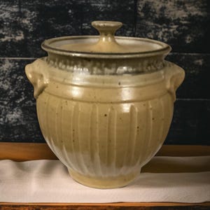 May include: A tan ceramic lidded pot with two side handles. The pot has vertical grooves and a rounded shape. The lid has a small knob. The pot is sitting on a white cloth on a wooden tray.