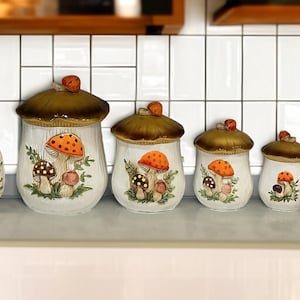 K&ouml;nnte beinhalten: Ein Set aus sechs Keramik-K&uuml;chendosen und einem Utensilienhalter mit wei&szlig;en K&ouml;rpern und bemalten orangefarbenen und braunen Pilzillustrationen. Jede Dose hat einen braunen pilzf&ouml;rmigen Deckel, w&auml;hrend der Utensilienhalter den Text KITCHEN UTENSILS zeigt.