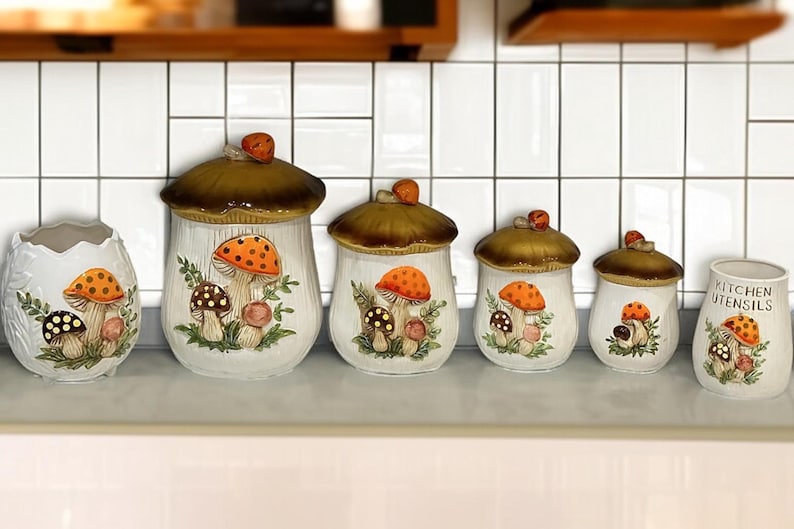K&ouml;nnte beinhalten: Ein Set aus sechs Keramik-K&uuml;chendosen und einem Utensilienhalter mit wei&szlig;en K&ouml;rpern und bemalten orangefarbenen und braunen Pilzillustrationen. Jede Dose hat einen braunen pilzf&ouml;rmigen Deckel, w&auml;hrend der Utensilienhalter den Text KITCHEN UTENSILS zeigt.
