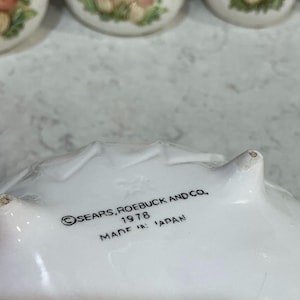 K&ouml;nnte beinhalten: Die Unterseite einer wei&szlig;en Keramikschale mit drei kleinen F&uuml;&szlig;en. Schwarzer Text auf der Unterseite lautet: SEARS, ROEBUCK AND CO., 1978, MADE IN JAPAN. Die Keramikoberfl&auml;che ist glatt und wei&szlig;, mit einer schwach gepr&auml;gten Sternmarkierung.