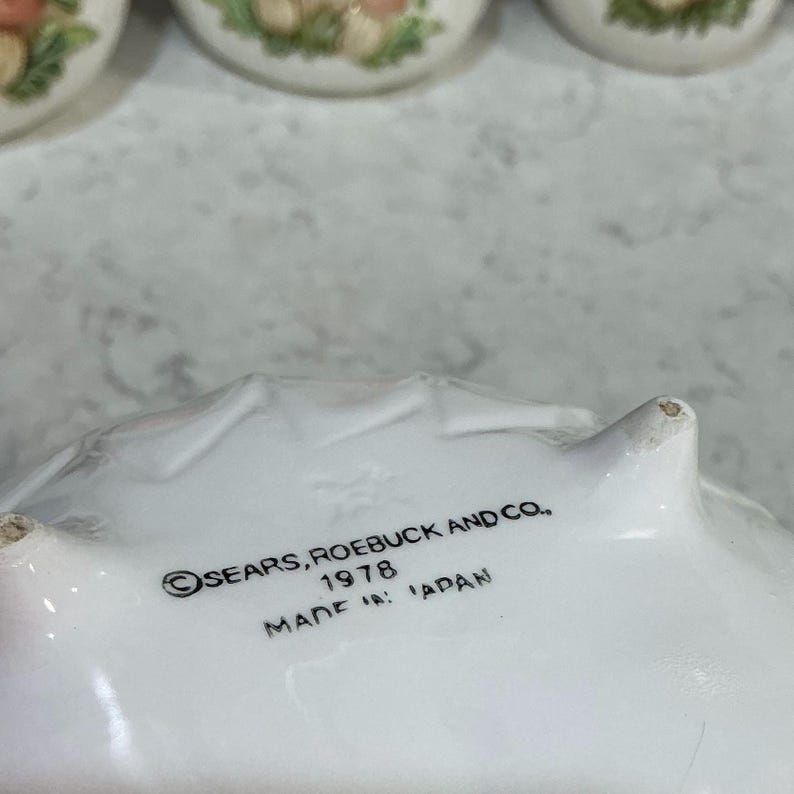 K&ouml;nnte beinhalten: Die Unterseite einer wei&szlig;en Keramikschale mit drei kleinen F&uuml;&szlig;en. Schwarzer Text auf der Unterseite lautet: SEARS, ROEBUCK AND CO., 1978, MADE IN JAPAN. Die Keramikoberfl&auml;che ist glatt und wei&szlig;, mit einer schwach gepr&auml;gten Sternmarkierung.