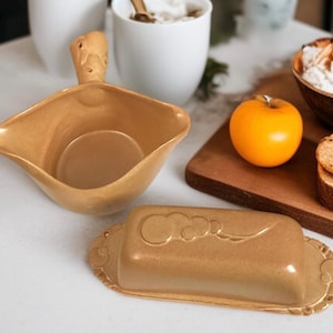 May include: A golden-colored butter dish and serving bowl are displayed on a white surface. The butter dish has a decorative cloud design on the lid. An orange apple and a wooden cutting board with bread are also in the image.