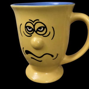 May include: Yellow ceramic mug with a blue rim and a hand-painted face with a grumpy expression. The face has two eyes, a nose, and a frowning mouth.
