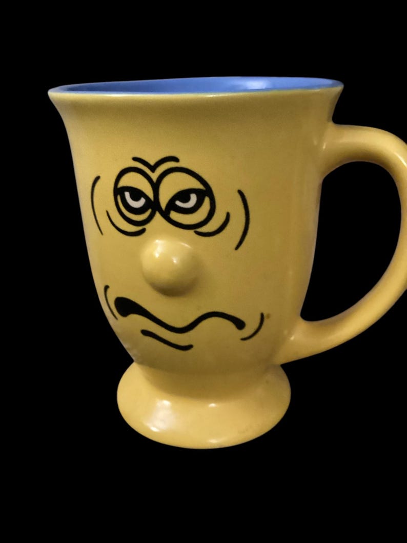 May include: Yellow ceramic mug with a blue rim and a hand-painted face with a grumpy expression. The face has two eyes, a nose, and a frowning mouth.