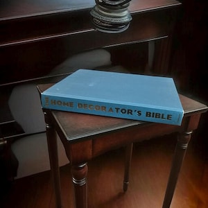 May include: A blue hardcover book titled "THE HOME DECORATOR'S BIBLE" rests on a small, dark wooden table. The book's title is in gold lettering. The table has four legs and is placed in front of a dark wooden cabinet.