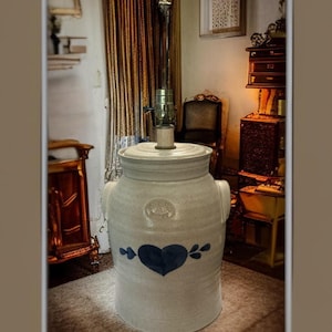 May include: A stoneware lamp base with a light blue glaze. The lamp features a dark blue heart design with leaf accents. The lamp has a cylindrical shape with a lid and two handles. The lamp is topped with a metal lamp fixture.