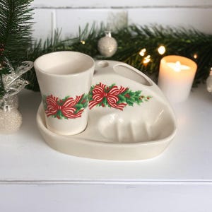 May include: Off-white ceramic toothbrush holder and cup set. The set features a red bow and holly design. The cup is cylindrical, and the holder has a soap dish and a slot for a toothbrush. Festive holiday decor.