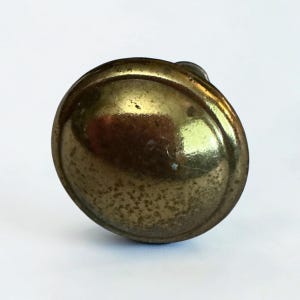 May include: A circular brass button with a domed, polished surface and a slightly weathered, textured patina. The button features a raised outer rim and a smooth, reflective center, showing signs of age and use across its metallic surface.