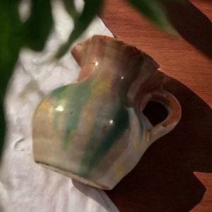 May include: A rustic, hand-painted ceramic pitcher with a brown, green, and yellow glaze. The pitcher has a wide, round body and a small, curved handle.