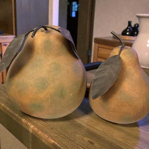 May include: Two decorative pear-shaped objects with attached leaves. The pears are primarily tan with green and brown spots, and the leaves are dark gray. They sit on a wooden surface, suggesting a rustic or farmhouse theme.