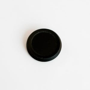 May include: A small, circular black object with a smooth, flat top surface and a slightly raised outer rim. The item features a solid matte black finish throughout, presenting a simple, uniform, and geometric design with a clean, circular silhouette.