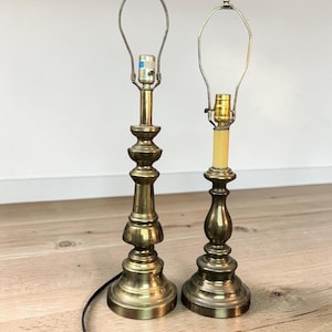 May include: Two brass table lamp bases with tiered, pedestal-style designs. Each lamp features a metal harp attachment at the top for a shade. The taller lamp has a silver-toned socket, while the shorter lamp has a gold-toned socket.