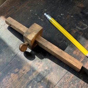 May include: A brown wooden marking gauge with a square beam and a circular tightening knob, positioned alongside a yellow wooden pencil with a silver ferrule and a pink eraser, showing a classic tool for woodworking layout and measurement.