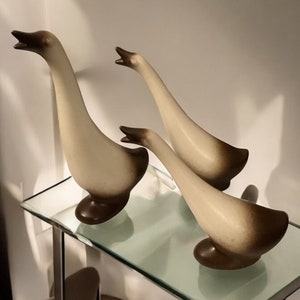 May include: Three ceramic goose figurines with a two-tone color scheme. The geese have a cream-colored body and a dark brown head and base. They are arranged on a glass shelf, with their necks arched upwards.