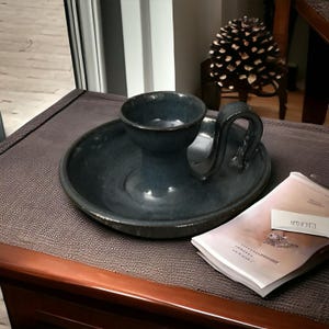 May include: A dark blue ceramic candle holder with a handle. The holder is on a brown surface with a book and a small white tag that says "Ella" on it.