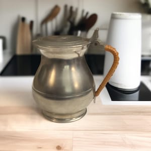 May include: A vintage pewter pitcher with a hinged lid and a woven handle. The pitcher has a bulbous body and a shiny, metallic finish. The handle is a light brown color. The pitcher is likely used for serving beverages.