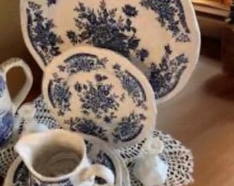 May include: A collection of white porcelain dishes with a blue floral pattern. The set includes a large oval platter, a smaller oval plate, a creamer, and several saucers. The dishes are arranged on a white lace doily.