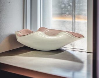 Könnte beinhalten: Dekorative Glasschale in Weiß und Rosa mit gewelltem Rand. Der Innenraum ist zartrosa, der Rand mit einem goldenen Akzent versehen. Die Schale steht auf einer neutralen Oberfläche in der Nähe eines Fensters.