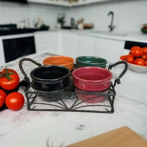 May include: A set of four small ceramic bowls in black, orange, green, and pink, held within a rectangular black wire metal caddy with two twisted handles. The bowls are arranged in a two-by-two grid inside the wire frame, which features a decorative base.