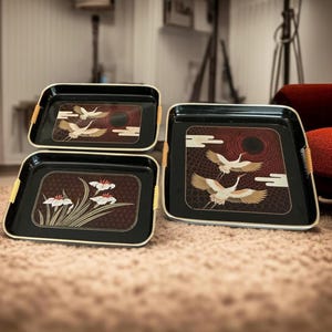 May include: Three rectangular black serving trays with rounded corners and light-colored woven trim on the side handles. Two trays feature illustrations of flying cranes, while the third tray displays a floral design with white petals.