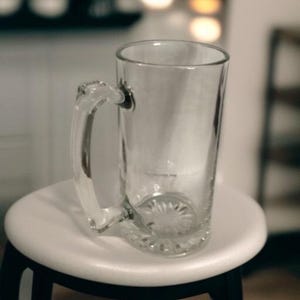 May include: A clear glass beer mug with a handle. The mug is empty and sits on a white stool. The handle is attached to the side of the mug. The base of the mug has a decorative pattern.