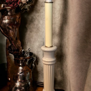 May include: A white ceramic table lamp with a cream colored shade and a gold trim. The lamp is on a wooden surface.