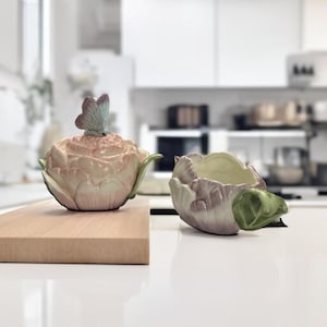 May include: A ceramic teapot and bowl set. The teapot is shaped like a flower with a butterfly on top, in shades of pink, green, and purple. The bowl is a matching color and design. The set is on a wooden cutting board.