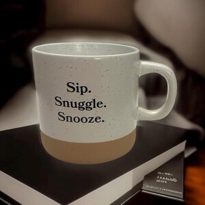 May include: White speckled ceramic mug with a tan base, featuring the words "Sip. Snuggle. Snooze." in black lettering. The mug has a curved handle and is resting on a stack of books.