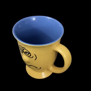 May include: Yellow ceramic mug with a blue interior and a cartoon face with a sad expression.