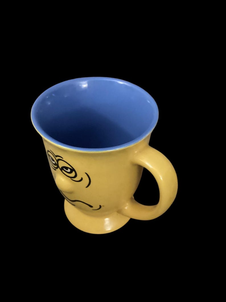 May include: Yellow ceramic mug with a blue interior and a cartoon face with a sad expression.