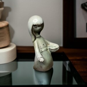 May include: A ceramic figurine of a stylized figure in a flowing robe. The figure has a rounded head with short bangs and a serene expression. The figurine is primarily a muted green color with darker accents and sits on a reflective surface.