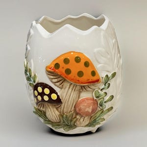 K&ouml;nnte beinhalten: Eine wei&szlig;e Keramikvase mit einem gezackten, unebenen oberen Rand. Die Au&szlig;enseite zeigt ein erhabenes, dreidimensionales Reliefdesign aus drei Pilzen in Orange, Braun und Rosa, akzentuiert durch gr&uuml;nes Blattwerk und gelbe Punkte.