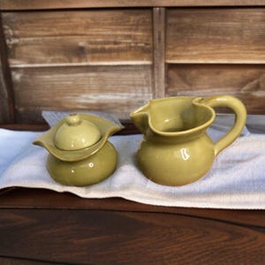 May include: A set of two ceramic sugar and creamer containers in a muted olive green color. The creamer has a unique, wavy design and a handle. The sugar container has a lid.