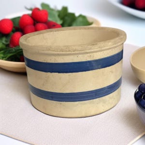 May include: A beige stoneware crock with two horizontal blue stripes. The antique-style container has a slightly oval opening and a textured surface. The crock is likely used for food storage or decoration. The background includes a wooden bowl with red berries.
