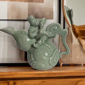 May include: A celadon green ceramic teapot with a sculpted figure riding atop. The teapot features a curved spout, a looped handle, and decorative swirling patterns. The figure is detailed with a hat and clothing.