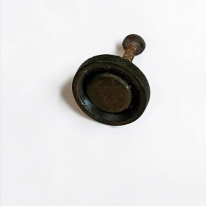 May include: A dark, circular metal knob with a recessed center and a threaded, rusted screw extending from the back. The hardware features a weathered, matte finish with visible signs of oxidation on the stem and a smooth, dark surface.