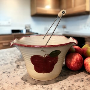 May include: A white ceramic apple-shaped bucket with a red apple design and a wire handle with a wooden knob. The bucket is sitting on a countertop with red apples beside it.