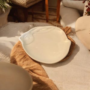 May include: A white ceramic plate shaped like a rabbit, with a smooth, glossy finish. The plate is resting on a brown and white fabric.