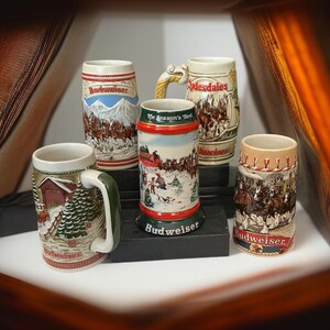 May include: A collection of vintage Budweiser beer steins. The steins feature various winter and holiday scenes, with the Budweiser logo prominently displayed. The steins are cylindrical with handles, and are made of ceramic. The steins are in various sizes and designs.