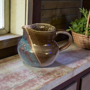 May include: A handcrafted ceramic pitcher with a brown and teal glaze sits on a distressed wooden surface. A small woven basket filled with green foliage is nearby. The scene is set near a window.