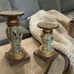 May include: Two decorative candle holders with a brown base and top. The body of the candle holders is painted in shades of green and gold, with leaf and floral designs. The candle holders are on a wooden surface.