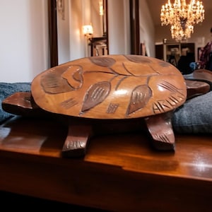 May include: A handcrafted wooden turtle-shaped table. The table has a detailed carved design on the shell, featuring leaf patterns. The turtle is brown and rests on four legs. The table is placed on a wooden surface.
