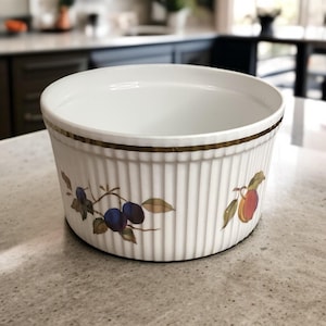 May include: White ceramic bowl with vertical ribbed design and a gold rim. The bowl is decorated with a plum and leaf motif. The bowl is sitting on a light gray countertop.