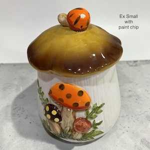 K&ouml;nnte beinhalten: Ein pilzf&ouml;rmiges Keramikgef&auml;&szlig; mit einem braunen Deckel und einem kleinen orangefarbenen Akzent. Die wei&szlig;e Basis zeigt erhabene, bemalte Pilz- und Blattmotive in Orange, Braun und Gr&uuml;n. Der Text auf der rechten Seite lautet: Ex Small with paint chip.