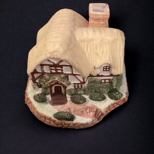 May include: A small ceramic cottage figurine with a thatched roof, brown trim, and green ivy details. The cottage has a chimney and a small door. The base of the figurine is decorated with green bushes and a stone path.