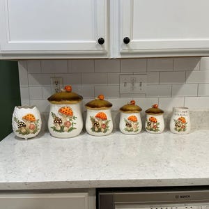 May include: A set of six ceramic kitchen canisters and containers featuring a white base with painted orange and brown mushroom designs. The collection includes four lidded jars, one open-top container, and one utensil holder labeled Kitchen Utensils.