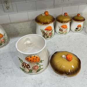 K&ouml;nnte beinhalten: Ein Set aus vier Keramik-K&uuml;chendosen mit wei&szlig;en K&ouml;rpern und braunen pilzf&ouml;rmigen Deckeln. Jede Dose zeigt ein erhabenes, handbemaltes Design aus orangefarbenen und braunen Pilzen mit gr&uuml;nem Blattwerk auf der Vorderseite und einem kleinen Pilzknauf.