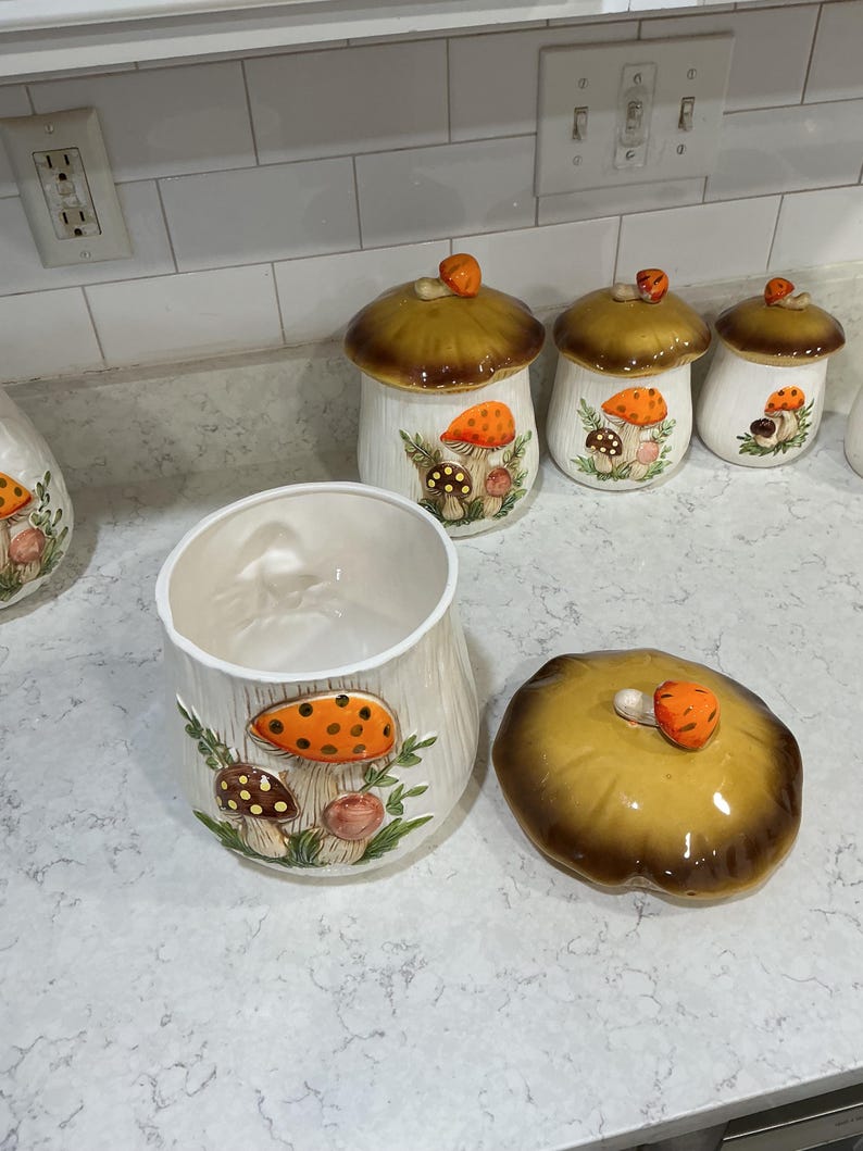 K&ouml;nnte beinhalten: Ein Set aus vier Keramik-K&uuml;chendosen mit wei&szlig;en K&ouml;rpern und braunen pilzf&ouml;rmigen Deckeln. Jede Dose zeigt ein erhabenes, handbemaltes Design aus orangefarbenen und braunen Pilzen mit gr&uuml;nem Blattwerk auf der Vorderseite und einem kleinen Pilzknauf.