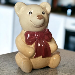 May include: A ceramic teddy bear figurine with a red bow. The bear is sitting with its paws in front of it and has a smiling face.